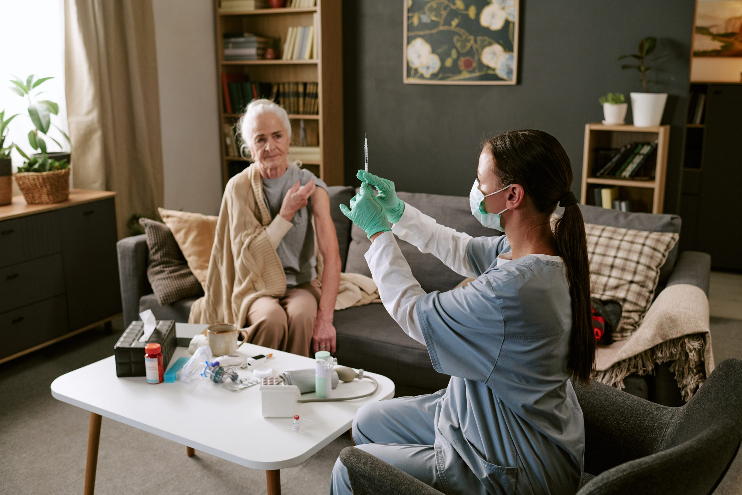 Senior Caucasian woman sitting on sofa preparing for vaccination while young adult Caucasian female nurse wearing mask and gloves holding syringe in living room setting