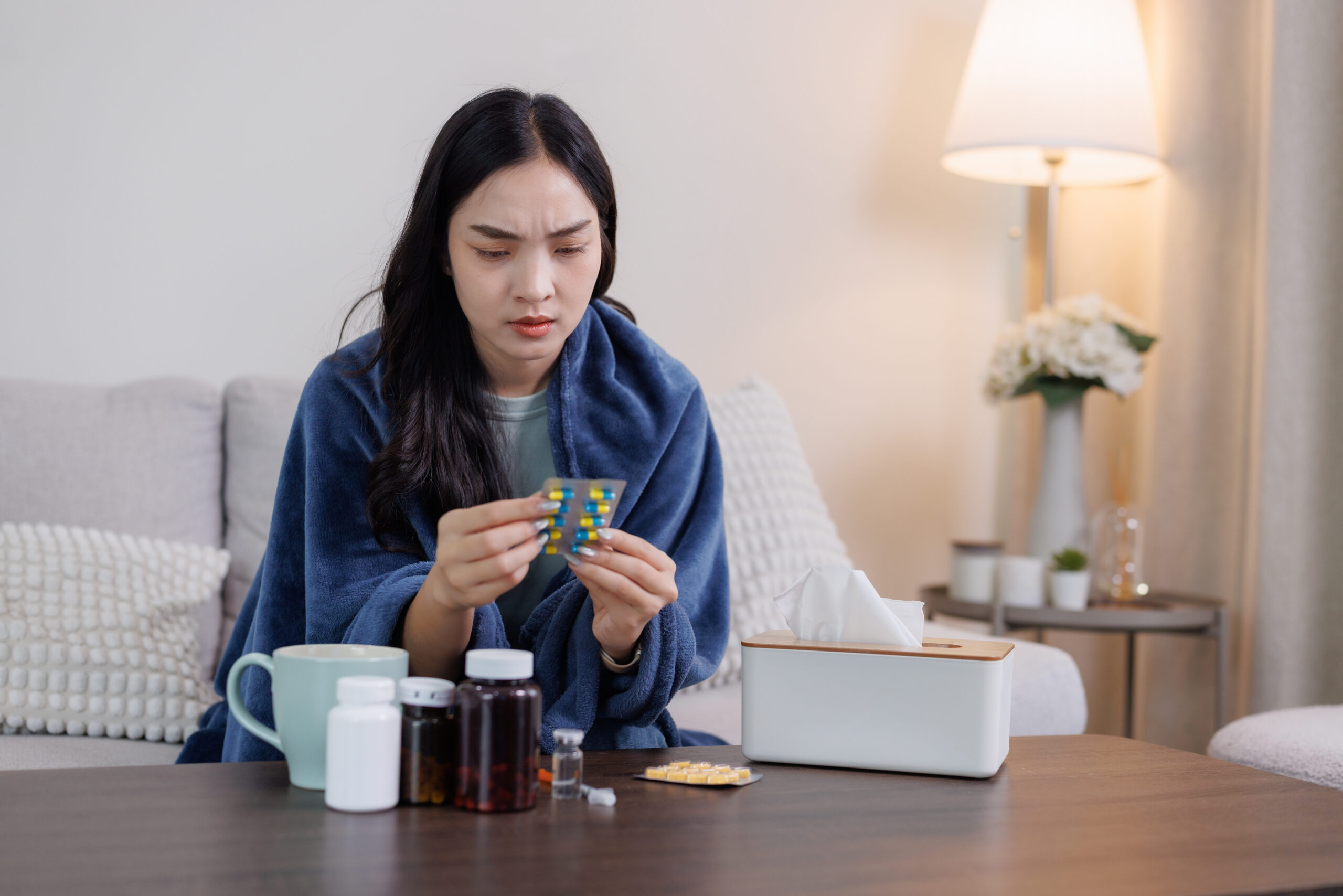 Woman feeling ill, wrapped in a blanket, looking at a blister pack of pills, with medicine bottles and tissues on a table
